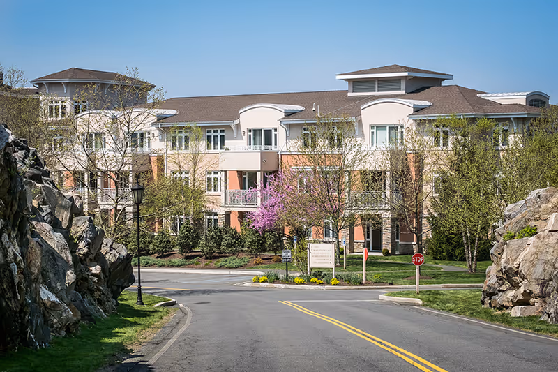 View of a multi-story residential building with balconies, surrounded by trees and landscaped greenery, with a paved road leading up to it and rocky formations on either side.