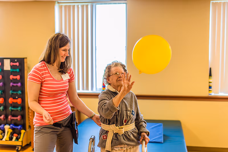 An elderly woman using a walker is playing with a yellow balloon while a female caregiver in a pink striped shirt stands beside her, smiling. They are in a room with exercise equipment and padded mats, with windows in the background.