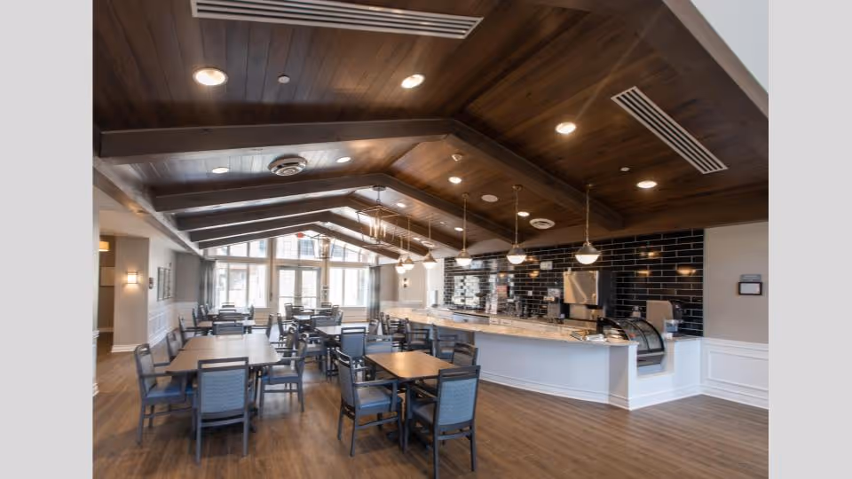 A spacious dining room with multiple tables and chairs arranged neatly. The room features a high wooden ceiling with exposed beams and recessed lighting. There is a long counter with pendant lights hanging above it and a black tiled backsplash behind the counter. Large windows at the far end allow natural light to fill the space.