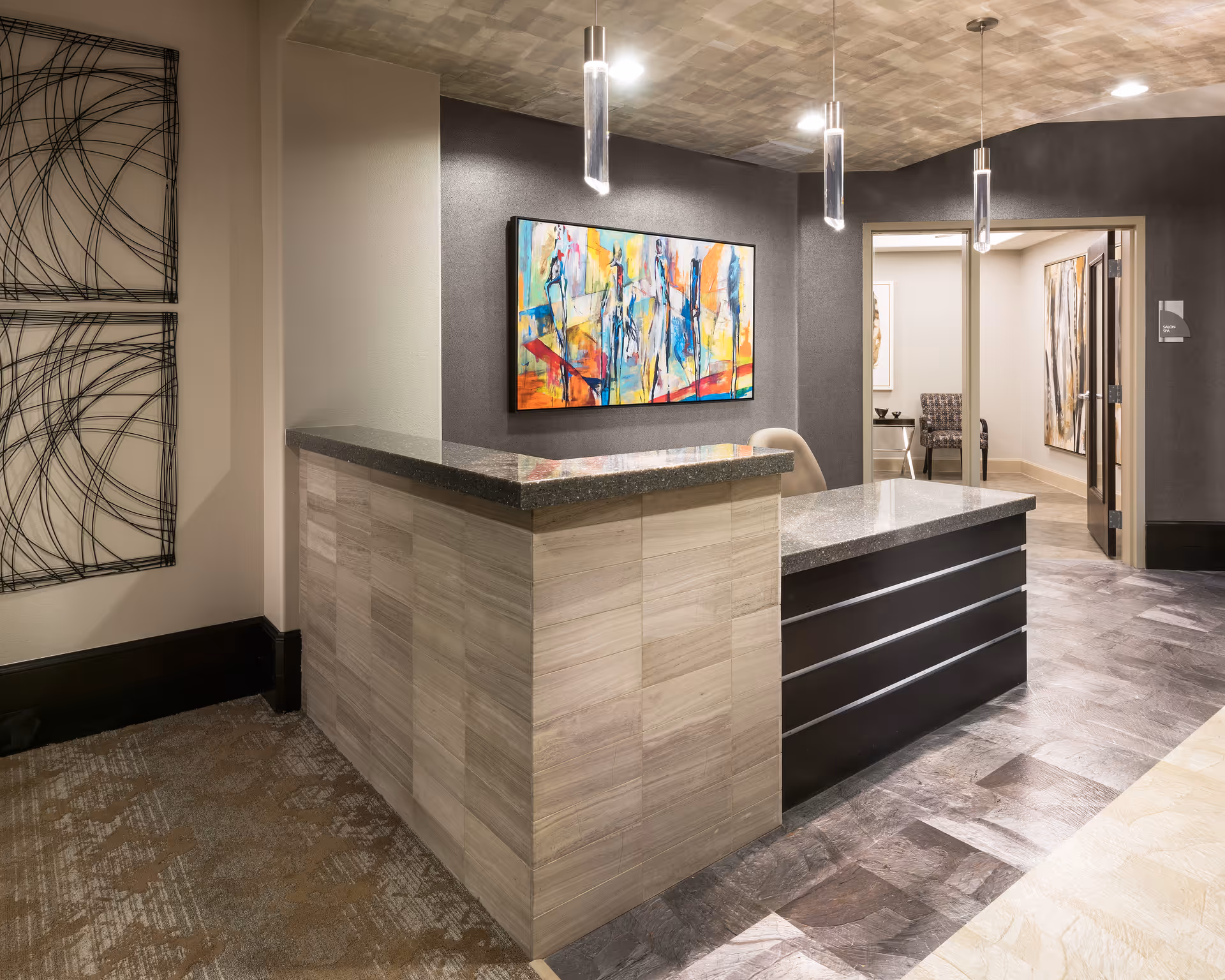 A modern reception desk area with a granite countertop and light wood paneling. Behind the desk is a colorful abstract painting on a dark gray wall. The ceiling has a textured finish with three pendant lights hanging above the desk. To the right, there is an open doorway leading to another room with additional artwork and chairs.