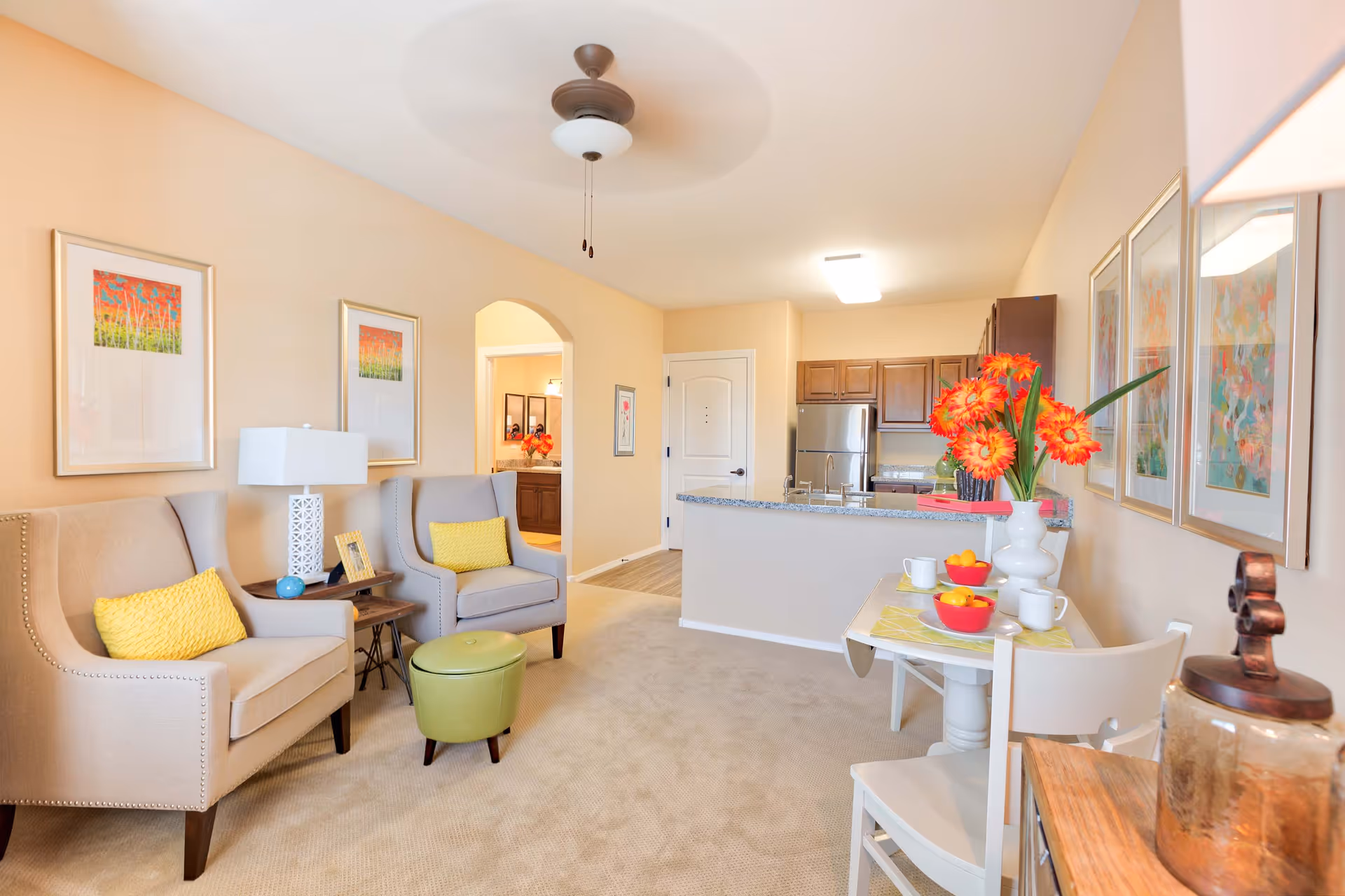A bright and cozy living area in a senior living facility featuring two beige armchairs with yellow cushions, a small green ottoman, a side table with a lamp and picture frame, and colorful artwork on the walls. The room opens into a kitchen with wooden cabinets, stainless steel appliances, and a granite countertop. A small dining table with white chairs is set with cups, bowls of fruit, and a vase of orange flowers.