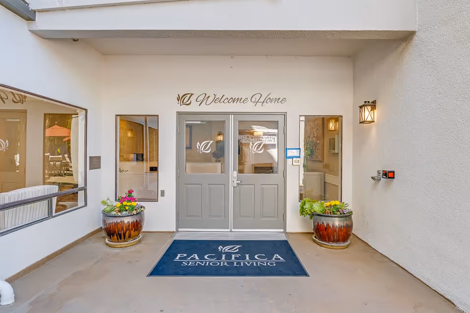 Entrance to a senior living facility with double gray doors, two large planters with flowers on either side, a welcome mat that reads 'Pacifica Senior Living', and a sign above the doors saying 'Welcome Home'.