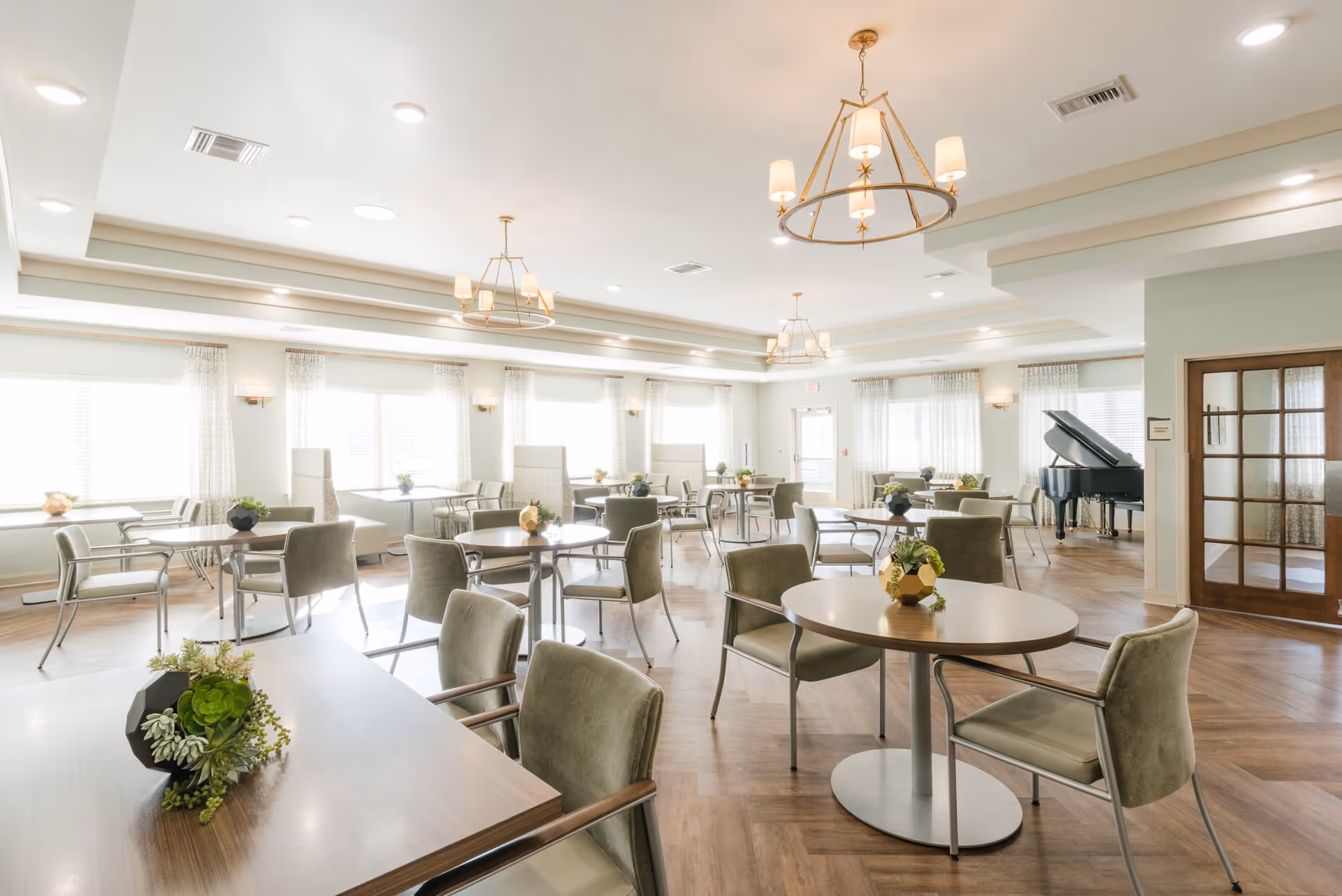 A bright and spacious dining room with multiple round and rectangular tables surrounded by cushioned chairs. The room features large windows with sheer curtains allowing natural light to fill the space. There are modern chandeliers hanging from the ceiling and a grand piano in the corner near a set of French doors.