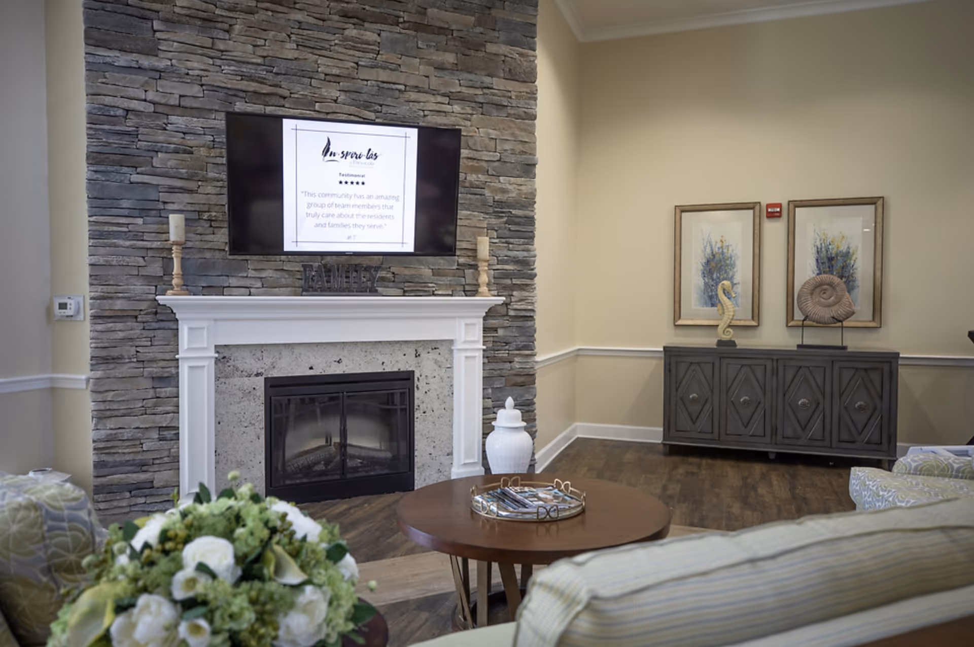 Communal sitting area featuring a stone fireplace with wall-mounted TV, round coffee table, floral arrangement, and a decorative sideboard with artwork.