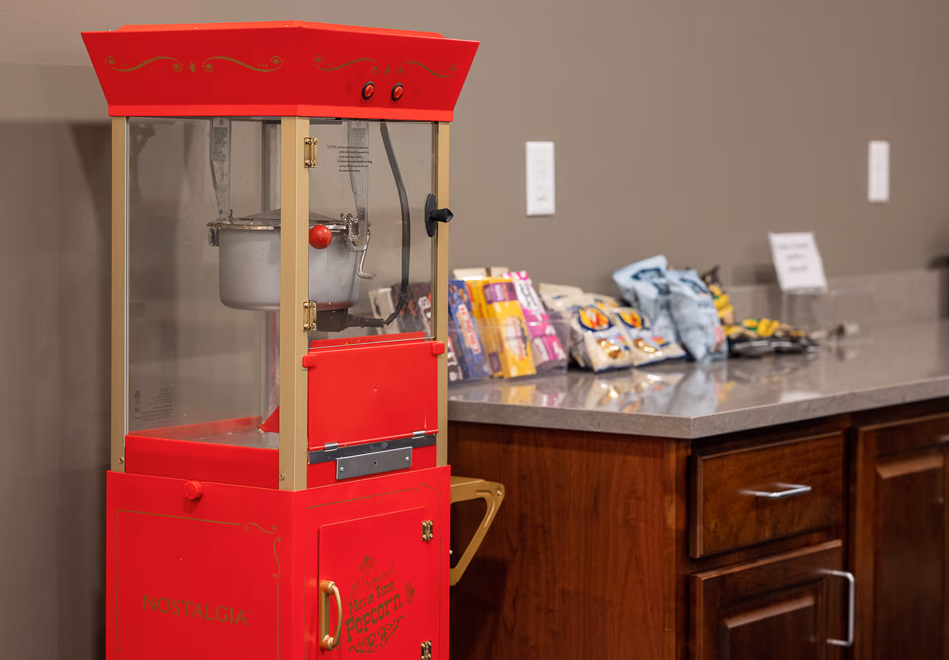 A red Nostalgia popcorn machine sits next to a countertop with snacks and wooden cabinets.