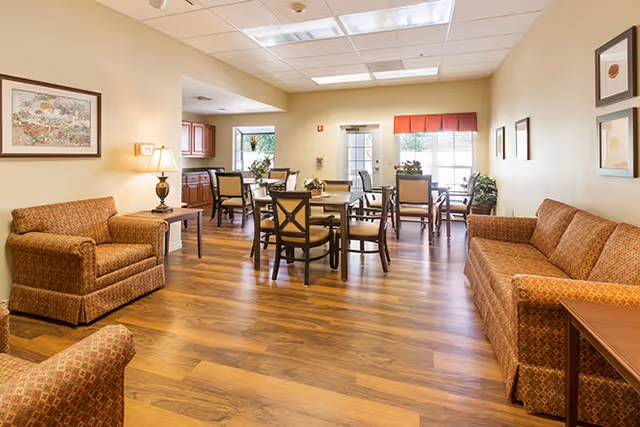 A well-lit common area in a senior living facility featuring wooden flooring, multiple tables with chairs, and comfortable upholstered seating including a sofa and armchairs. The walls are decorated with framed artwork, and there is a lamp on a side table. A window with a red valance allows natural light into the room.