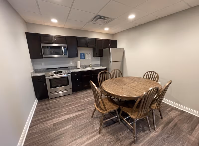 A kitchen area with dark wood cabinets, a stainless steel microwave, stove, and refrigerator. There is a round wooden dining table with six matching wooden chairs on a wood-patterned floor. The walls are light-colored, and the ceiling has recessed lighting.