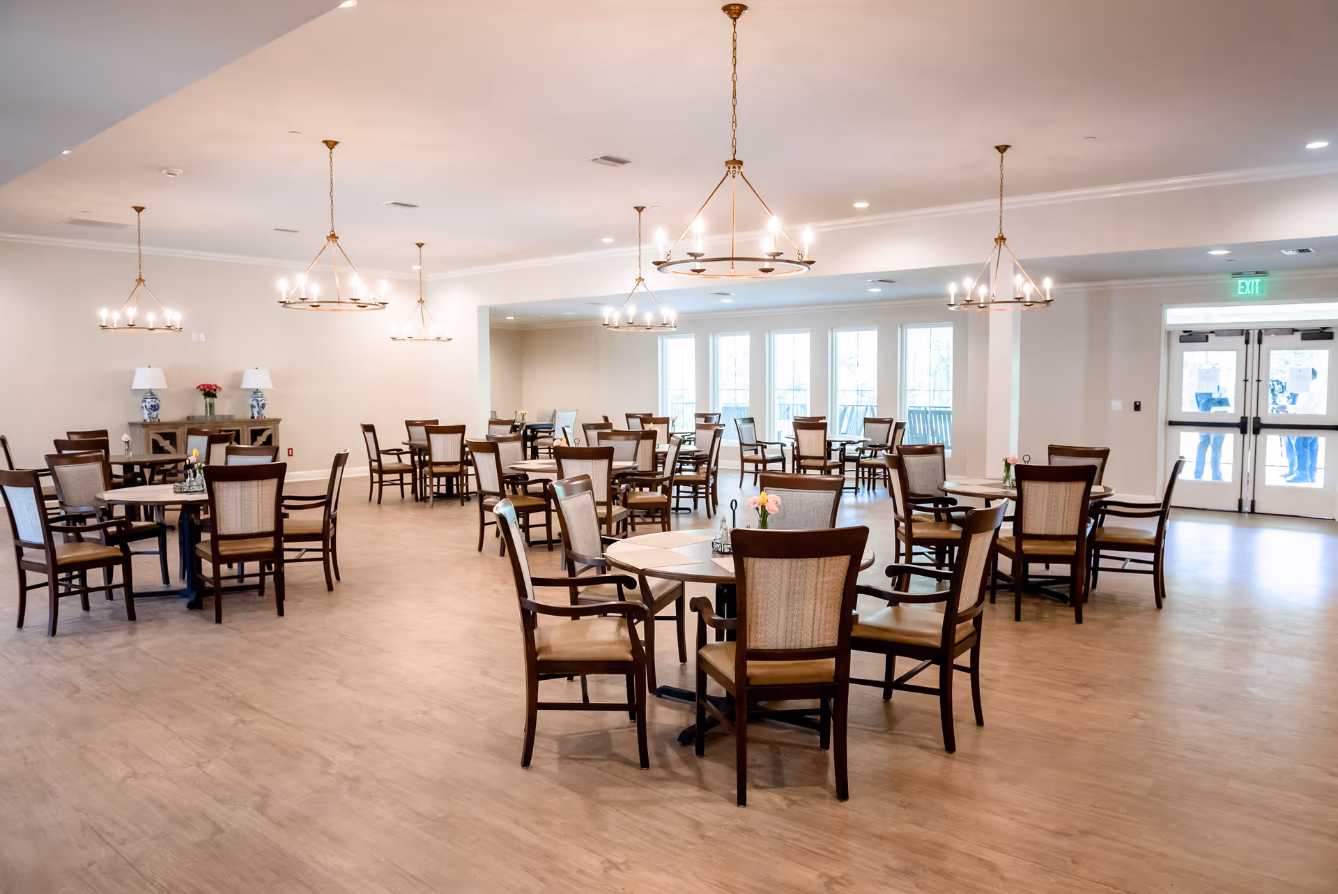 A spacious dining room with multiple wooden tables and chairs arranged neatly. The room has light-colored walls and wooden flooring, with several modern chandeliers hanging from the ceiling. There are large windows and glass doors letting in natural light, and a sideboard with two lamps and a flower vase is visible against one wall.