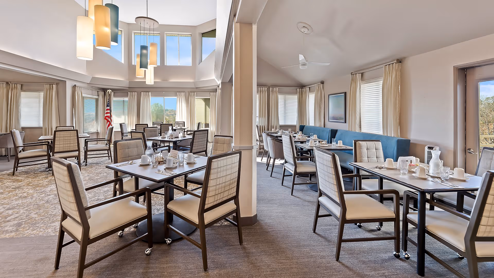 A bright and spacious dining room with multiple tables and chairs arranged neatly. The room features large windows with beige curtains allowing natural light to fill the space. There are modern pendant lights hanging from the ceiling and an American flag is visible near the windows. The tables are set with cups, glasses, and pitchers, ready for dining.