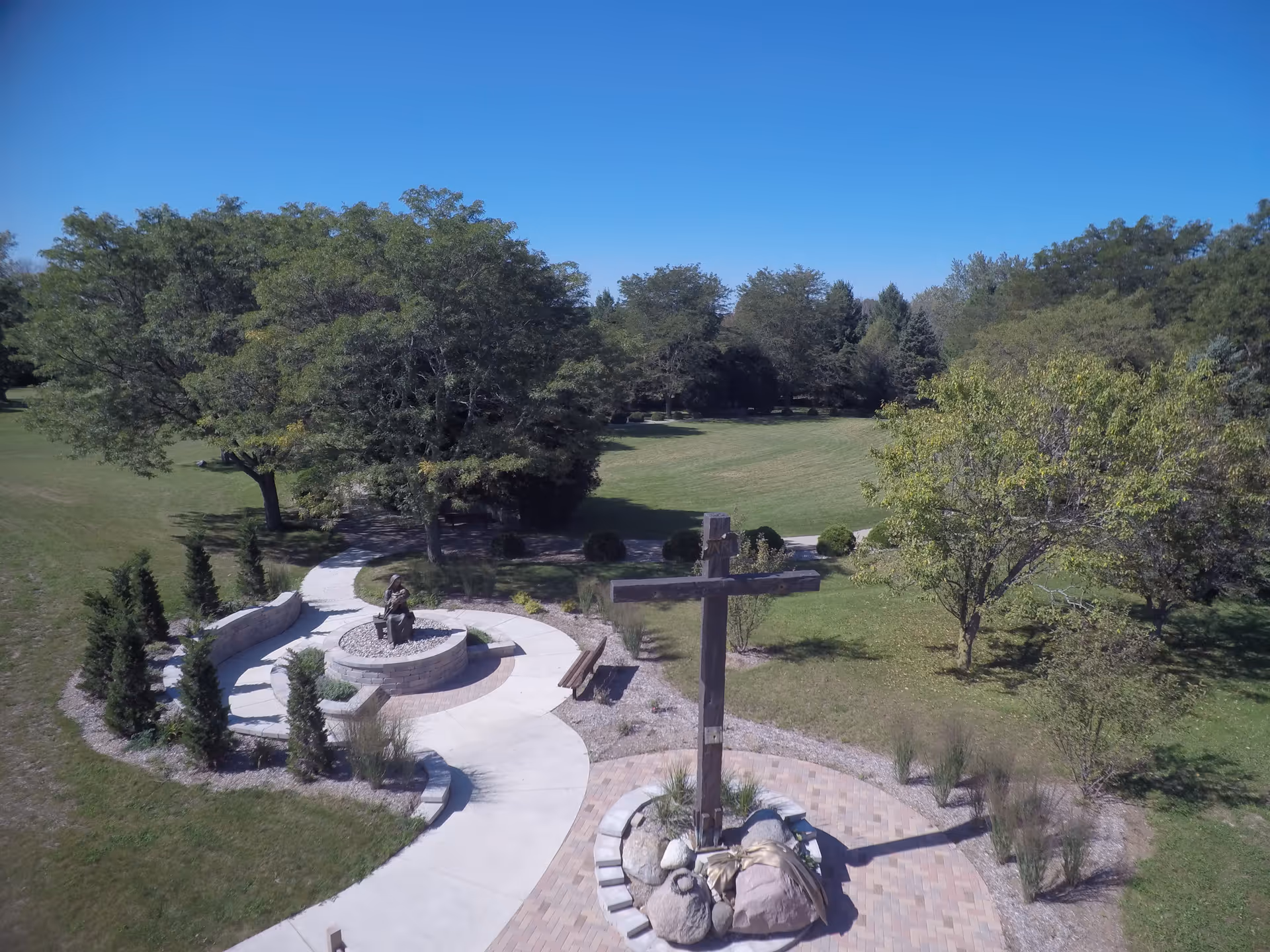 Outdoor garden area with a large wooden cross surrounded by rocks and plants, a curved paved walkway, a circular stone seating area with a statue, and several trees under a clear blue sky.