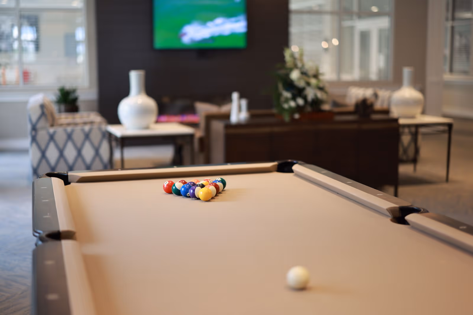 A beige pool table with a full rack of colorful billiard balls and a white cue ball in the foreground, set in a cozy lounge area with patterned armchairs, white vases on side tables, a dark wood console with a floral arrangement, and a TV mounted on the wall in the background.