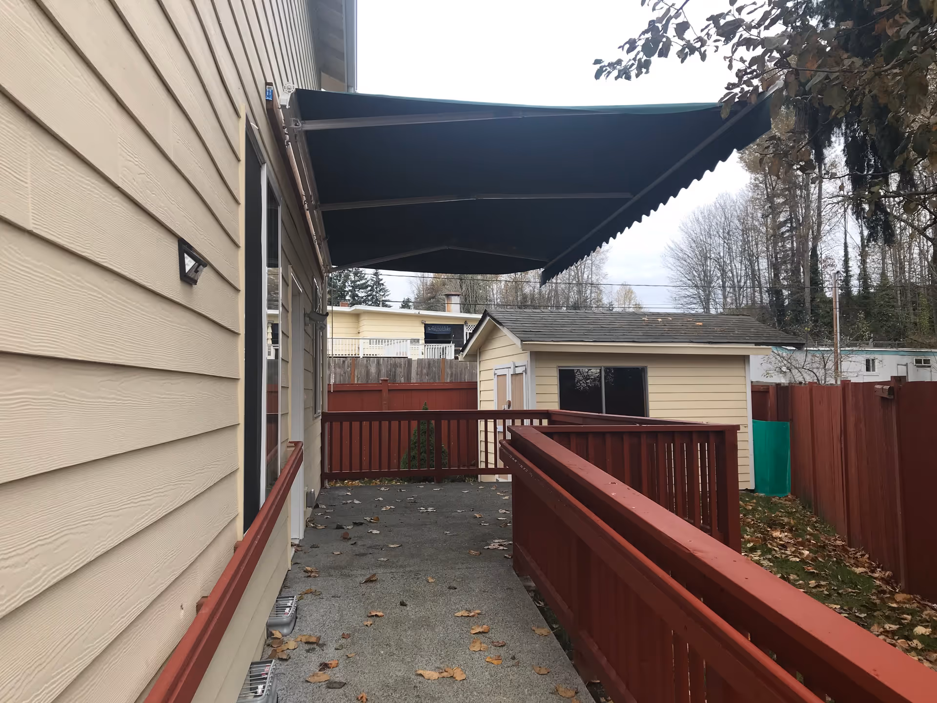 Outdoor view of a beige building with a red wooden railing and a concrete pathway covered with fallen leaves. A black awning extends from the building over the pathway. In the background, there is a small beige shed with a window, a red wooden fence, and trees with bare branches.