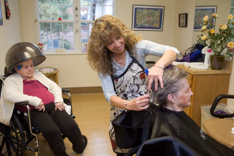 A woman hairstylist is cutting the hair of an elderly woman seated in a chair, while another elderly woman in a wheelchair sits nearby under a hair dryer in a well-lit room with windows and framed pictures on the wall.