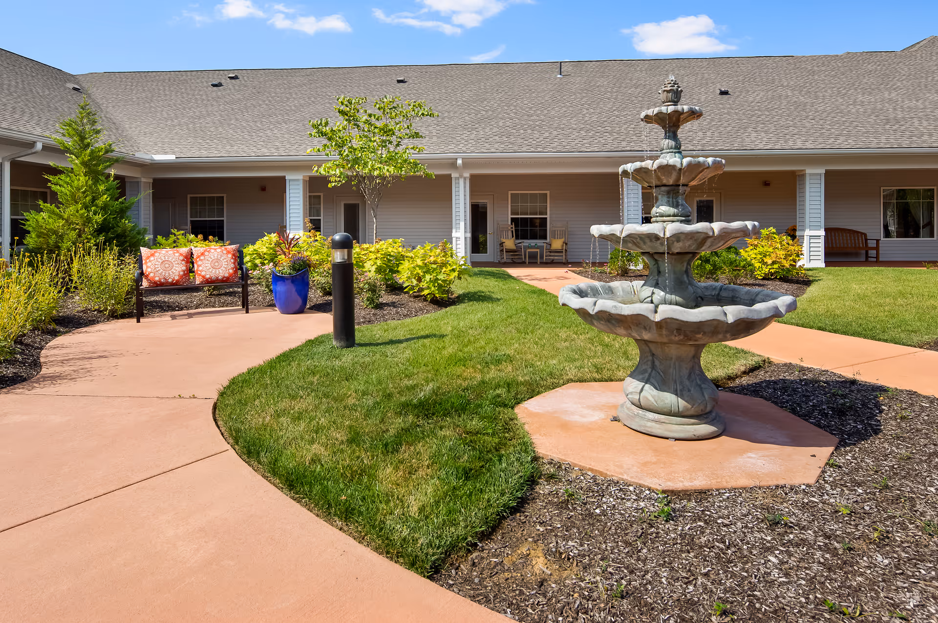 Outdoor courtyard area of a senior living facility with a three-tiered stone fountain, green grass, a curved concrete walkway, a bench with orange patterned cushions, potted plants, and a building with a covered porch and rocking chairs in the background under a blue sky with some clouds.