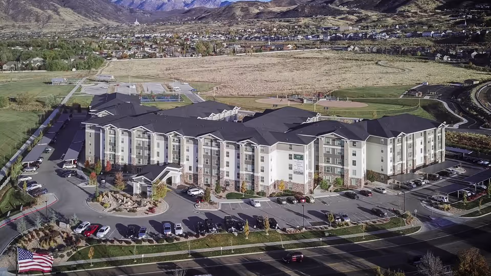 Aerial view of a large multi-story senior living building with surrounding parking, landscaped grounds, nearby sports fields, and mountains in the background.