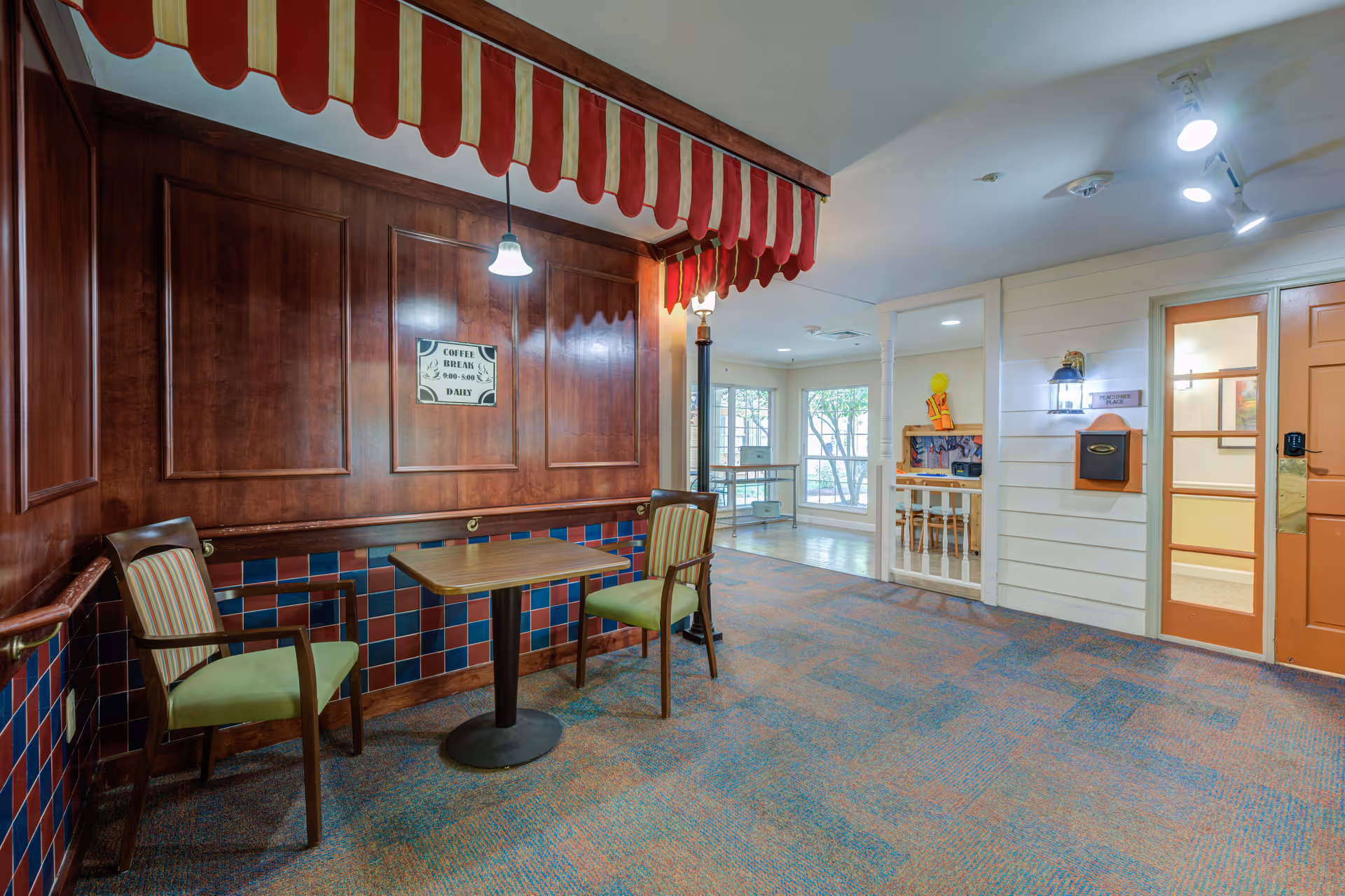 Indoor seating area with two wooden chairs with green cushions and a small wooden table between them. The area features a red and yellow striped canopy overhead and wood-paneled walls with a sign that reads 'Coffee Break 9:00-9:30 Daily.' The floor is carpeted with a multicolored pattern. In the background, there is a hallway with large windows letting in natural light and a door with a glass panel.