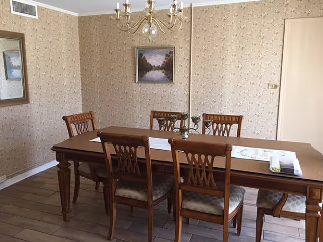 A dining room with a wooden dining table and six matching chairs. The table has a white runner and some decorative items on it. The walls are covered with beige floral wallpaper, and there is a framed painting of a landscape hanging on the wall. A chandelier with multiple lights hangs from the ceiling.