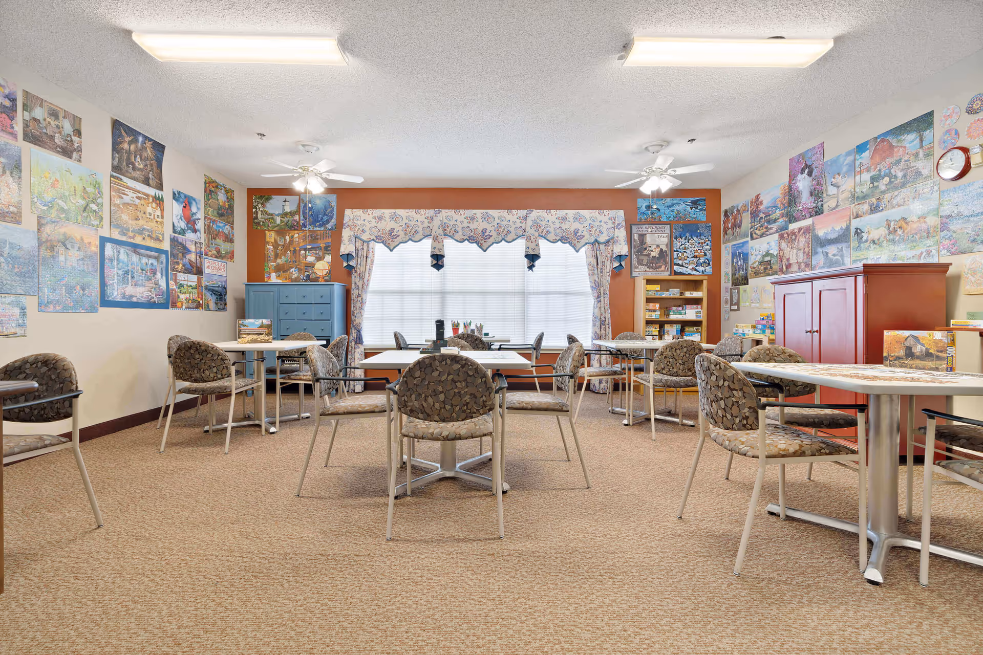 A cozy activity room featuring several tables and chairs, adorned with colorful artwork on the walls. The room has a large window with decorative curtains, a blue cabinet, and a red storage unit filled with games and supplies.