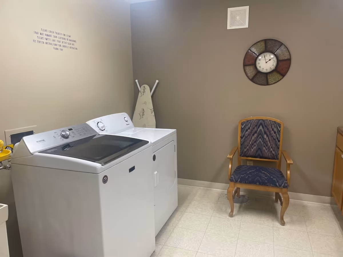 Laundry room with a white washing machine and dryer side by side, an ironing board leaning against the wall, a wooden chair with patterned upholstery, a decorative wall clock, and a beige wall with a small vent near the ceiling.