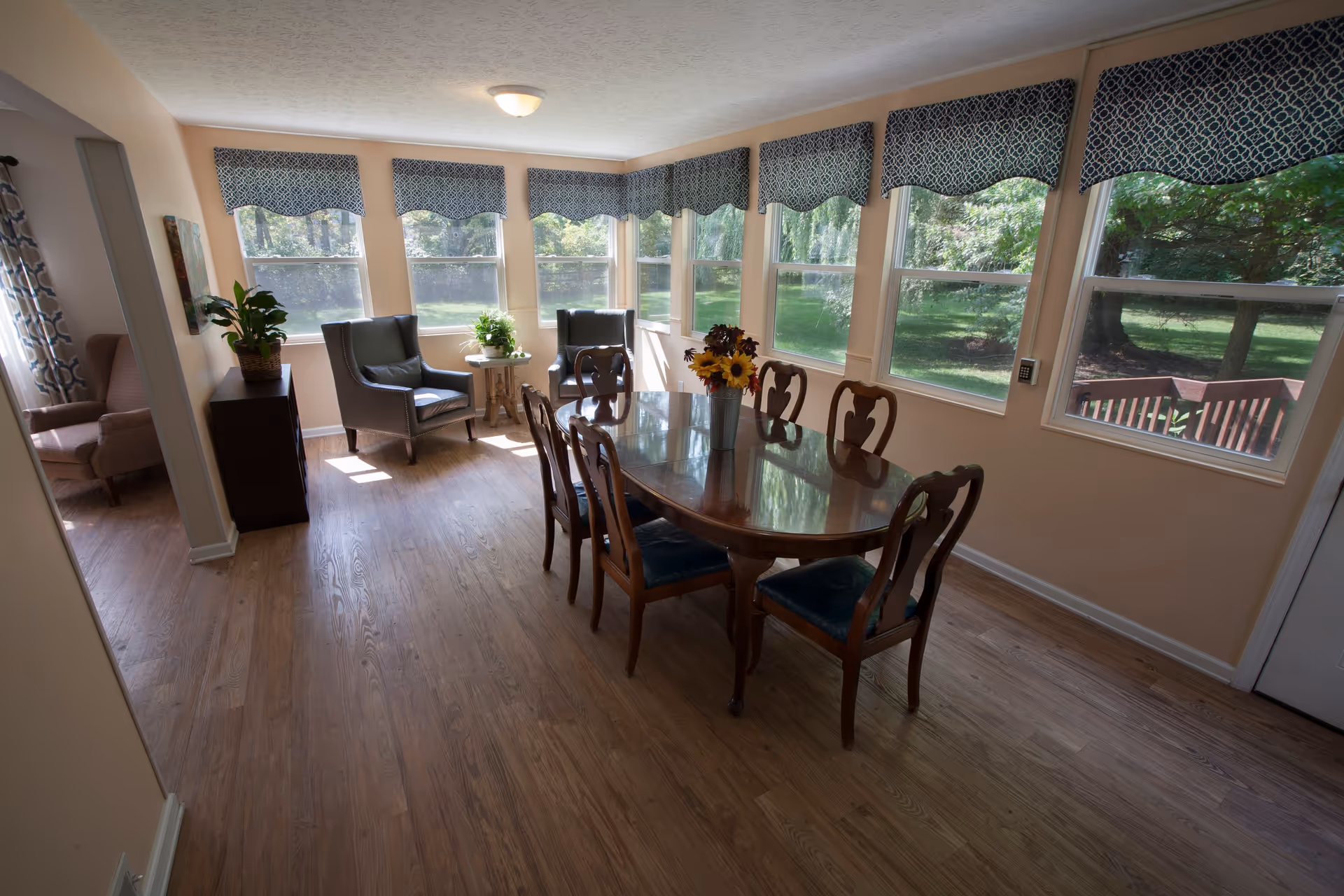 Sunlit dining room with a wooden table and chairs and armchairs by a row of large windows overlooking a green yard.