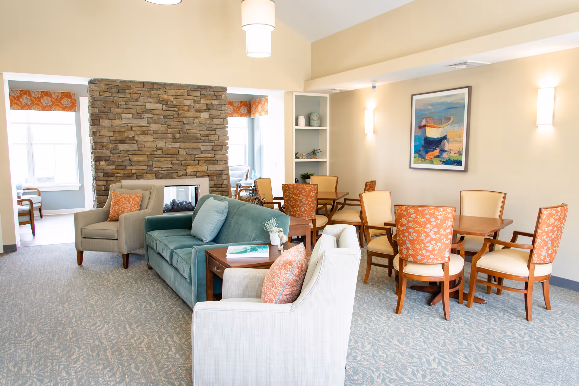A bright and spacious common area in a senior living facility featuring a stone fireplace, a teal sofa, beige armchairs with orange patterned pillows, and wooden tables with chairs that have orange floral upholstery. The room has carpeted flooring, soft wall lighting, and a colorful abstract painting on the wall.