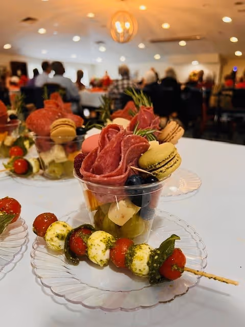 Close-up of plated appetizers—plastic cups with salami, macarons, fruit and cheese, plus caprese skewers—on a table at a dining event with guests blurred in the background.