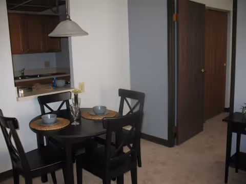 Small dining area with a dark round table set with place settings, four chairs, a kitchen pass-through, and a hallway door.