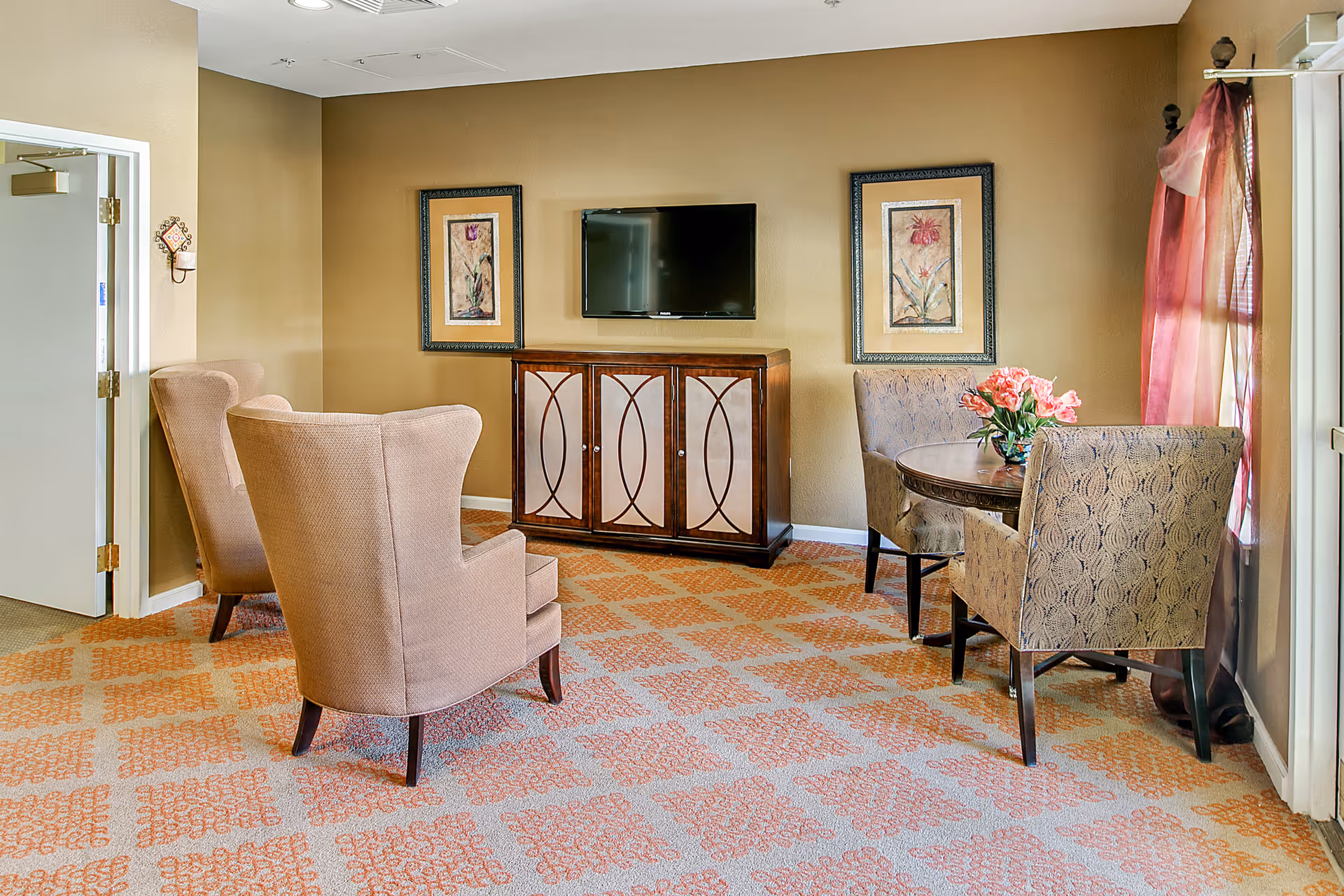 A cozy living room area with two beige armchairs facing a wooden cabinet with a flat-screen TV mounted above it. To the right, there is a round table with a flower vase and two patterned chairs near a window with sheer pink curtains. The room has beige walls and a patterned carpet.