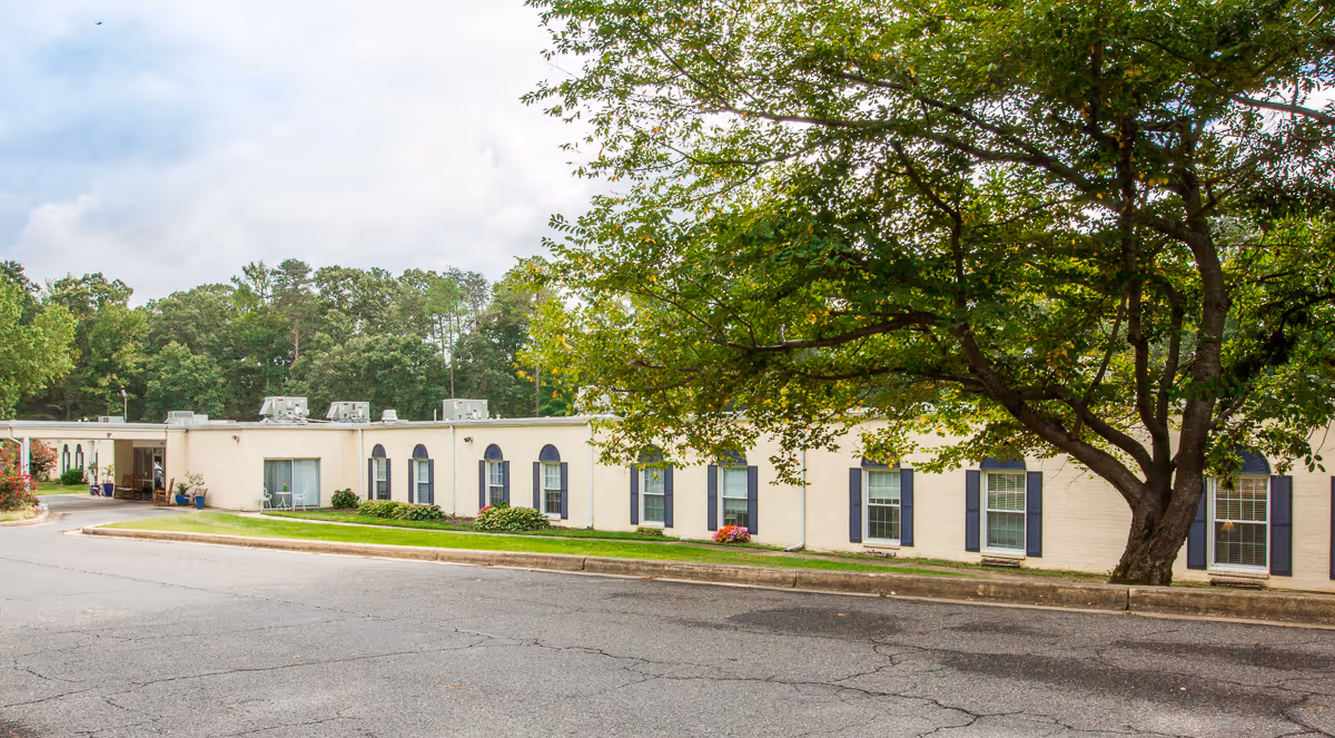 Exterior view of a single-story nursing center building with multiple windows, a large tree in the foreground, and a paved driveway in front. The building is surrounded by greenery and trees in the background under a partly cloudy sky.