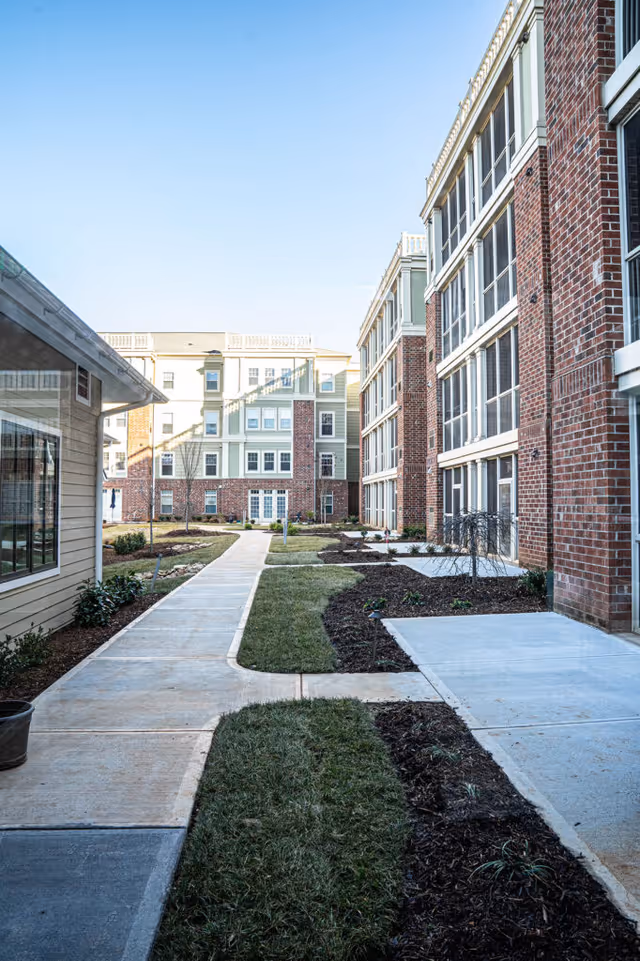 Outdoor walkway between buildings at The Woodlands at Furman, featuring brick and siding exteriors, landscaped grass and mulch areas, and clear blue sky.