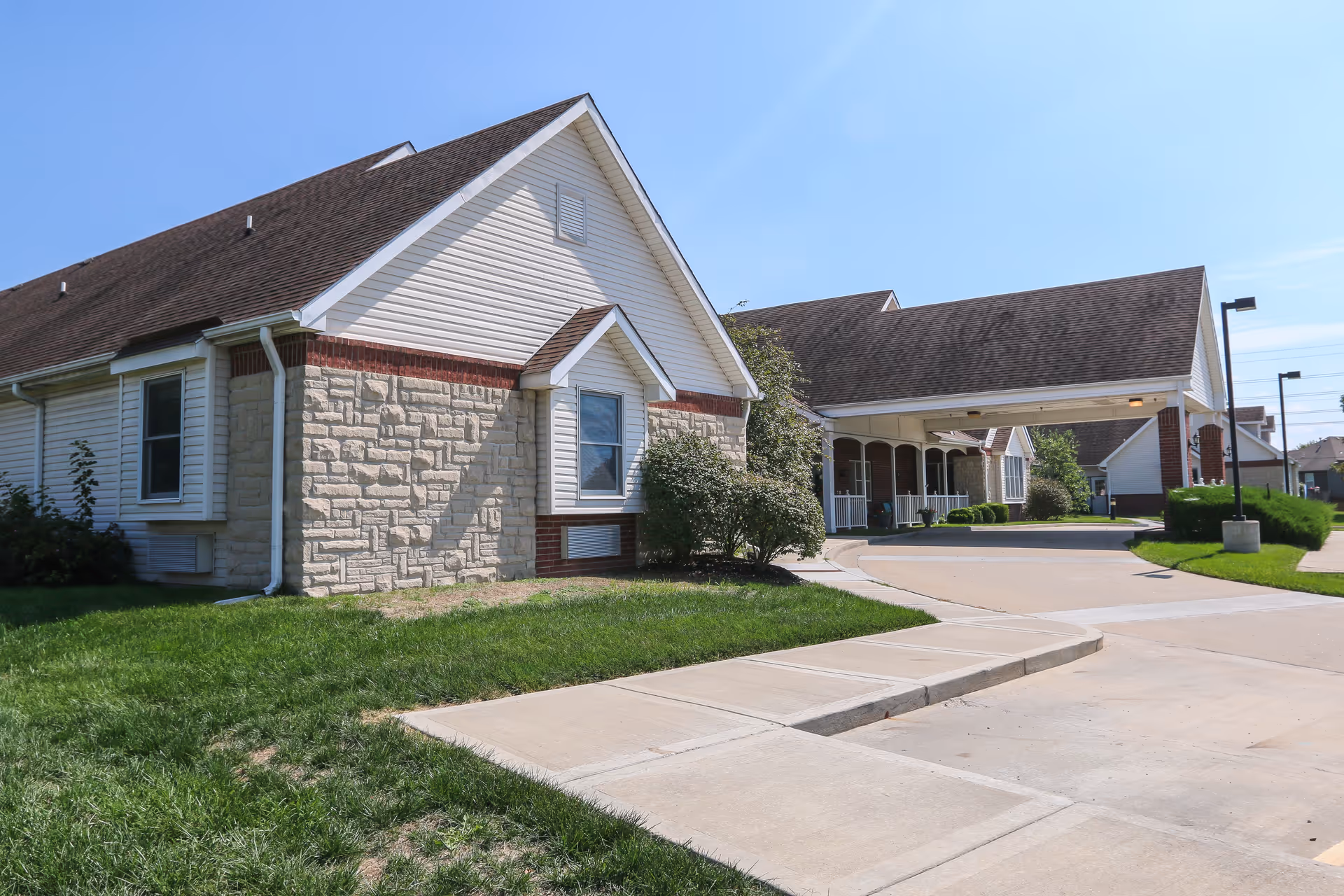 Exterior view of a senior living facility building with beige stone and white siding, a covered entrance driveway, green lawn, and clear blue sky.