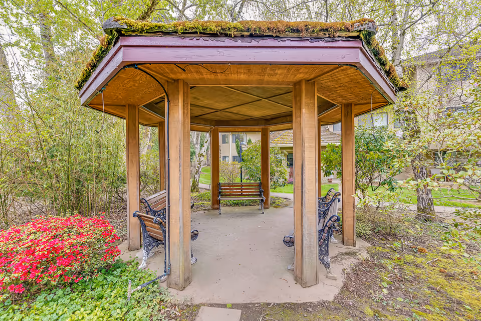 Wooden octagonal gazebo with benches in a landscaped courtyard surrounded by greenery and residential buildings.