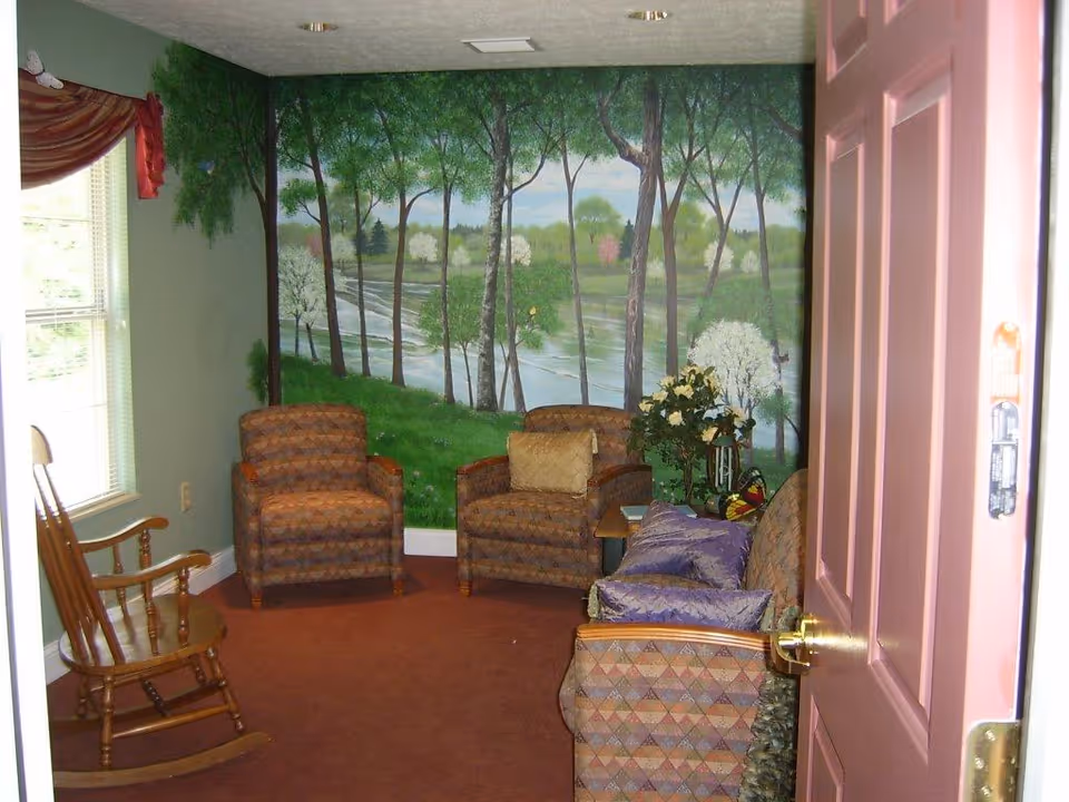 Small sitting room with patterned armchairs, a wooden rocking chair, a floral arrangement, and a mural of trees and a river on the back wall.