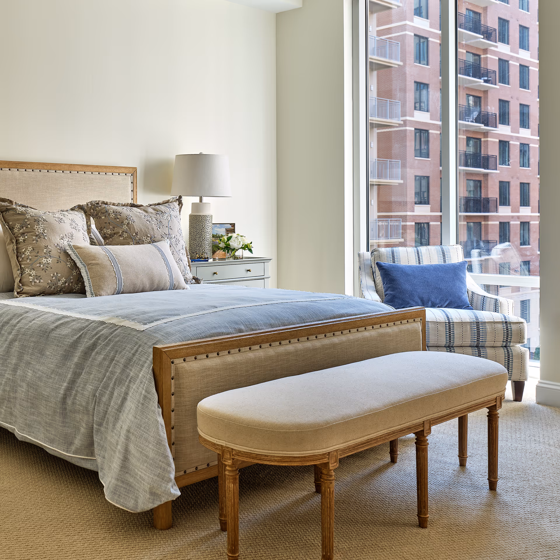 A bright bedroom with a neatly made bed featuring floral and solid pillows, a wooden bench at the foot of the bed, a bedside table with a lamp and flowers, and a striped armchair with a blue cushion next to a large window showing an exterior view of a brick building with balconies.