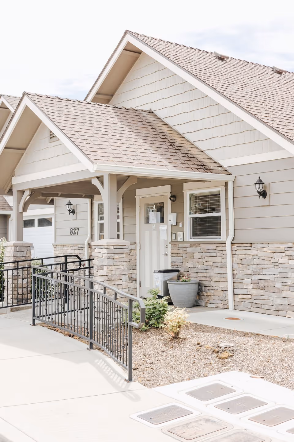Exterior view of a residential building entrance with a covered porch, stone and siding exterior walls, a ramp with black metal railing, and a potted plant near the door.