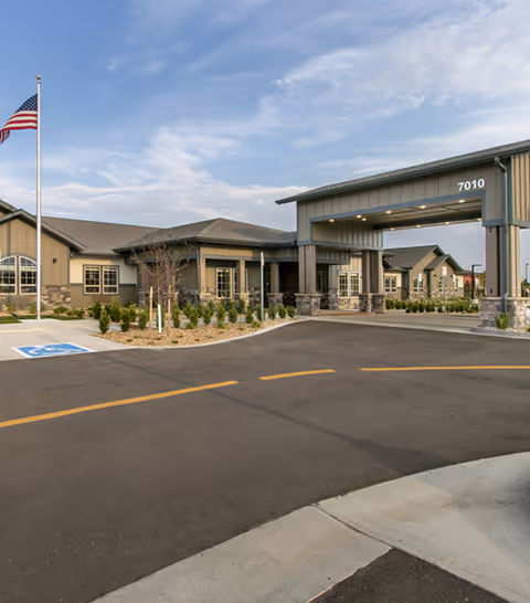 Exterior view of a single-story senior living facility building with a covered entrance driveway, landscaped bushes, an American flag on a flagpole, and a clear sky with some clouds.