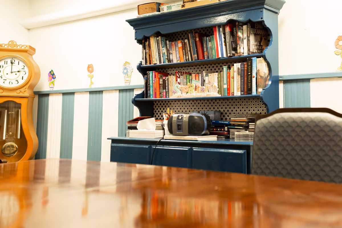 A cozy interior common room with a blue bookcase filled with books and a radio, a grandfather clock, striped wainscoting, and a polished table in the foreground.