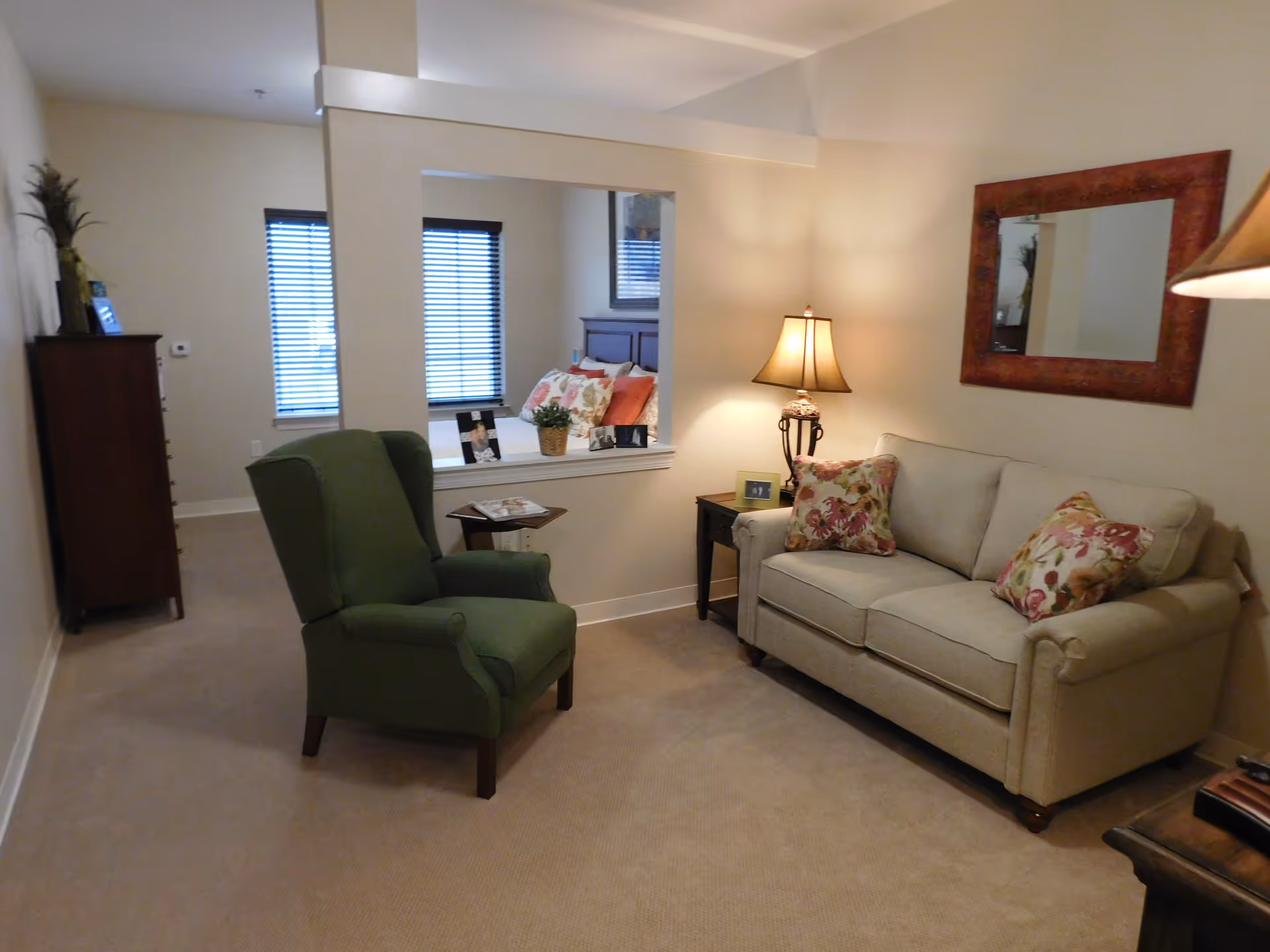 A cozy living room area with a beige sofa adorned with floral pillows, a green armchair, a side table with a lamp, and a large mirror on the wall. Through an open wall space, a bedroom with a bed, pillows, and two windows with blinds is visible.