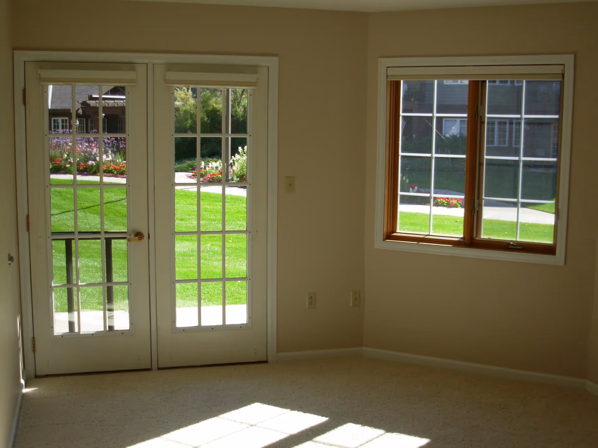 Empty bright room with beige walls, carpet, French doors and a window looking out onto a green lawn and garden.