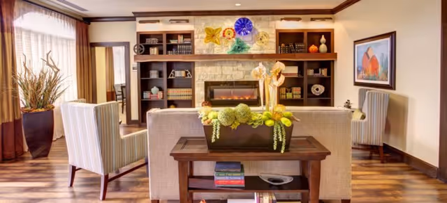 A cozy living room with a beige sofa and two striped armchairs facing a stone fireplace with built-in wooden shelves filled with books and decorative items. A wooden table behind the sofa holds a planter with green and yellow flowers. The room has wooden flooring, a large window with sheer curtains, and a framed painting on the wall.
