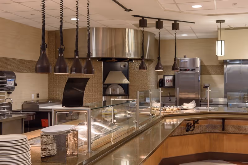 Interior view of a commercial kitchen area with a serving counter, glass sneeze guard, hanging pendant lights, stainless steel appliances including ovens and refrigerators, and stacks of plates on the counter.