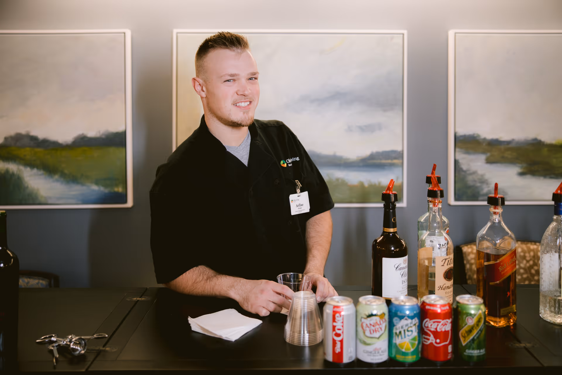 A man wearing a black shirt with a name tag stands behind a bar counter with various bottles of alcohol and soda cans in front of him. Behind him are three landscape paintings featuring green fields and cloudy skies.