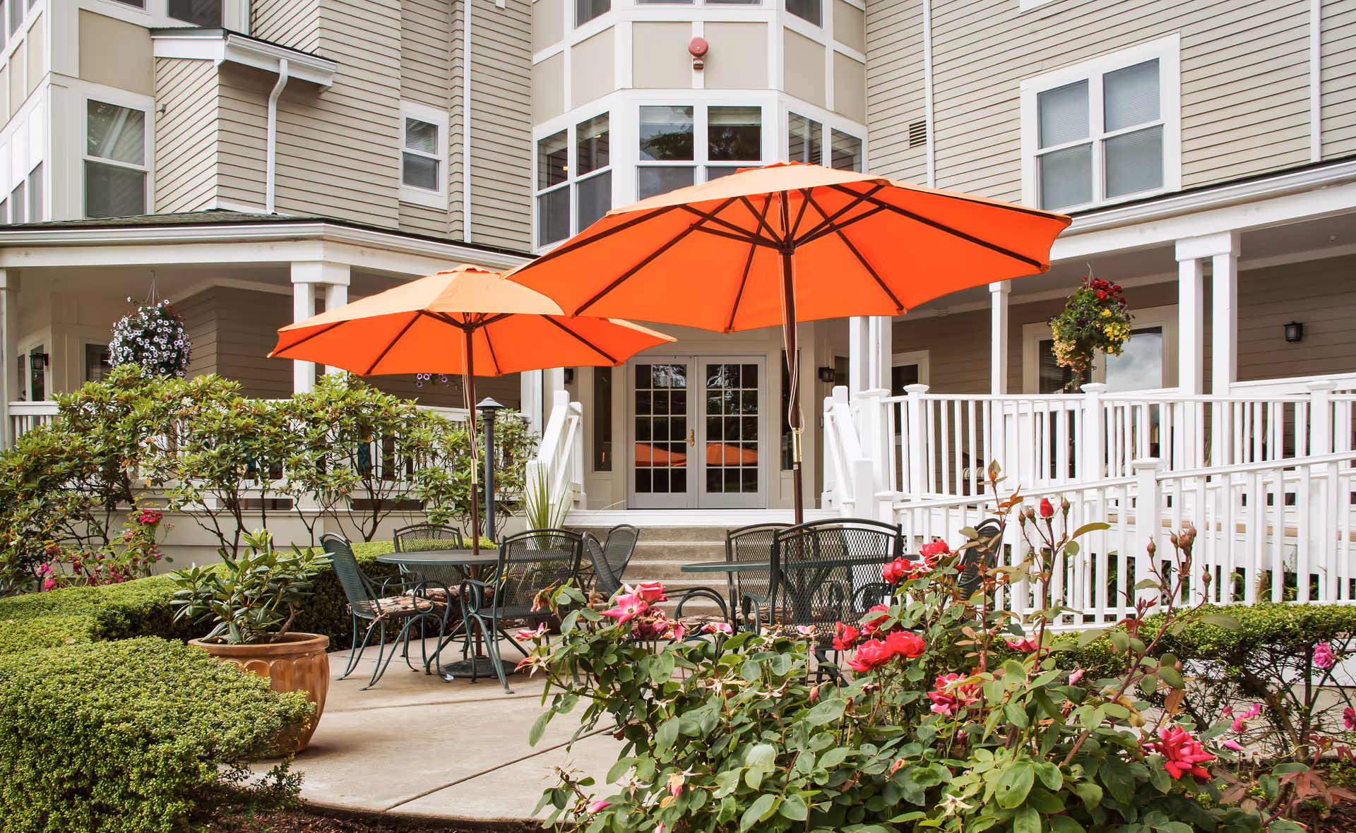 Outdoor patio area at Vineyard Park of Mercer Island featuring two orange umbrellas over metal tables and chairs, surrounded by green bushes and blooming pink flowers, with the building's beige exterior and white railings in the background.