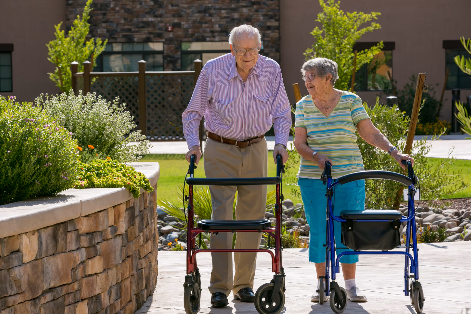 An elderly man and woman walking outdoors on a paved path using walkers. The man is wearing a striped shirt and beige pants, and the woman is wearing a striped shirt and blue pants. They are surrounded by greenery and plants with a building in the background.