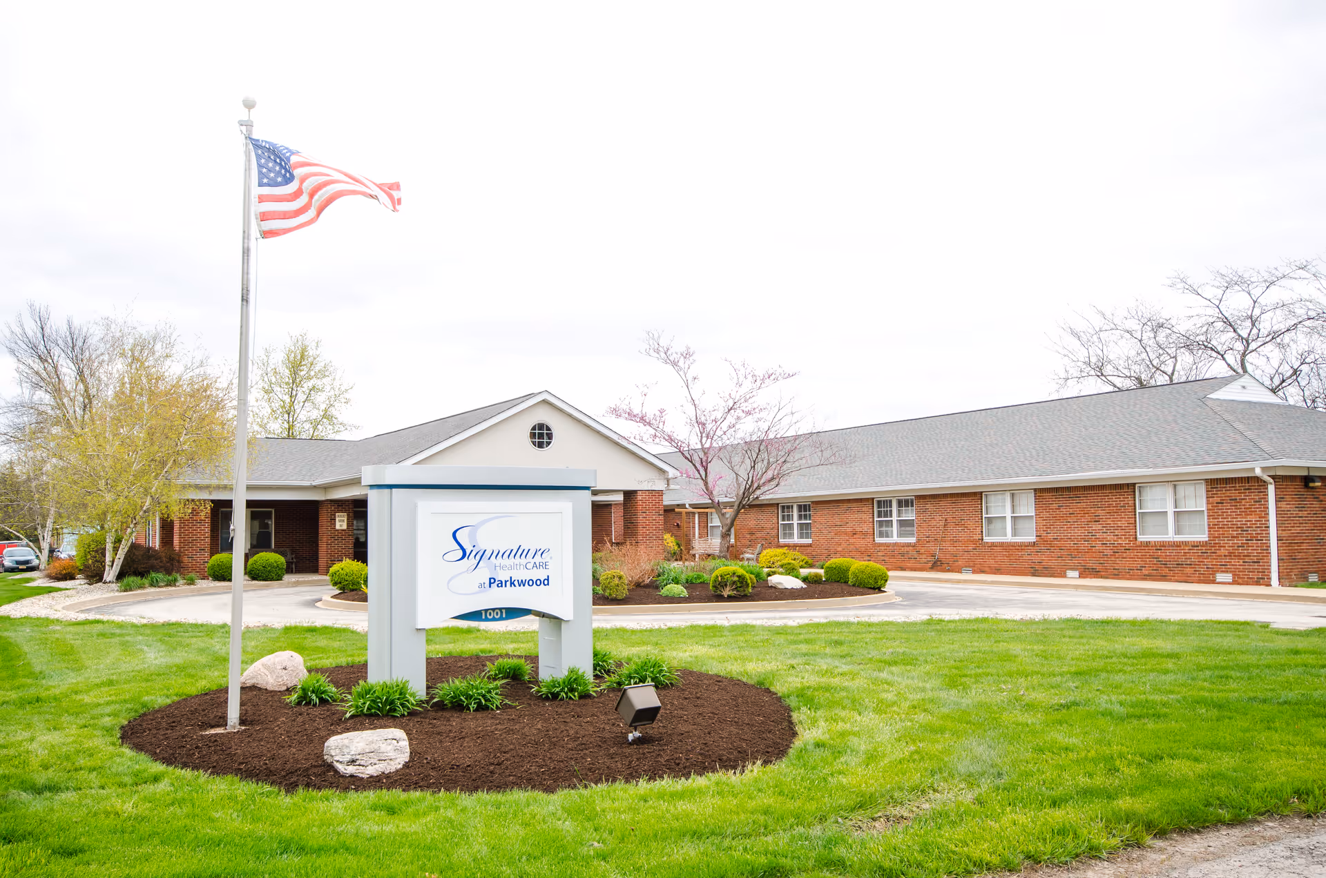 Exterior view of Signature HealthCARE at Parkwood building with a green lawn, a flagpole flying the American flag, and a sign displaying the facility's name and address.