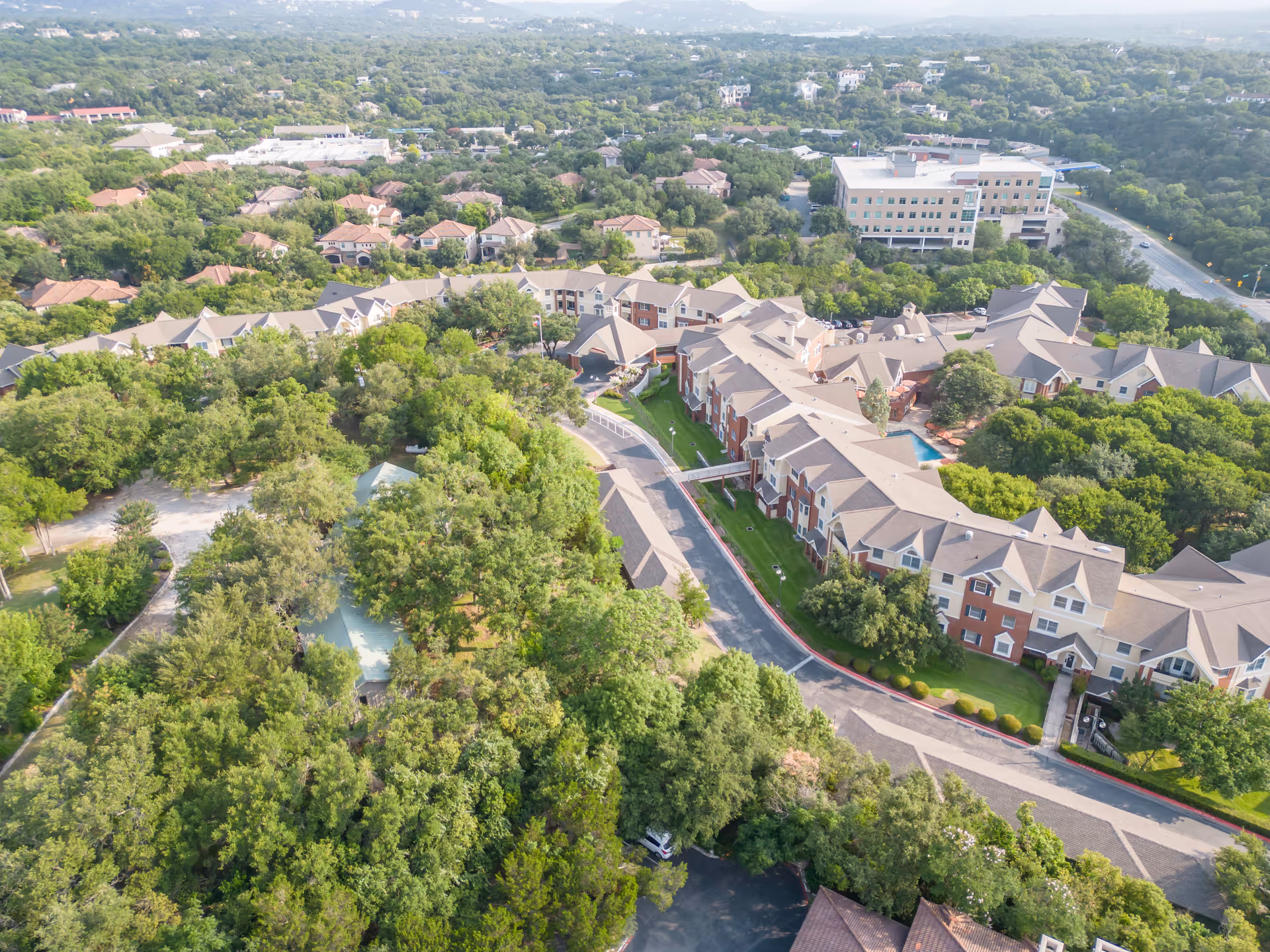 Aerial view of Brookdale Westlake Hills senior living facility surrounded by dense green trees and residential houses, with a large multi-story building and a road visible in the background.