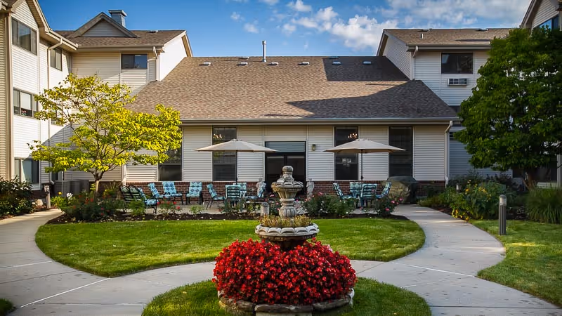 Courtyard with a central stone fountain surrounded by red flowers, curved walkways, patio seating with umbrellas, and the multi-story building facade.