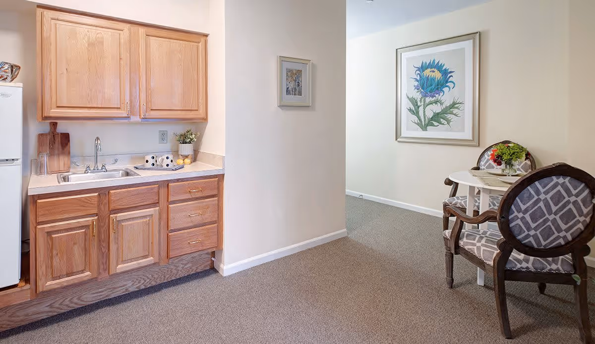 A small kitchen area with wooden cabinets, a sink, and a white refrigerator on the left. To the right, there is a small round table with two upholstered chairs featuring a geometric pattern. The walls are light-colored and decorated with framed floral artwork.