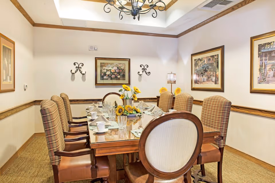 A dining room with a long wooden table set for a meal, featuring eight upholstered chairs with plaid backs and leather seats. The table is decorated with a centerpiece of sunflowers and neatly arranged tableware including cups, glasses, and napkins. The room has cream-colored walls with wooden trim, framed floral and street scene paintings, and a decorative chandelier hanging from a tray ceiling.