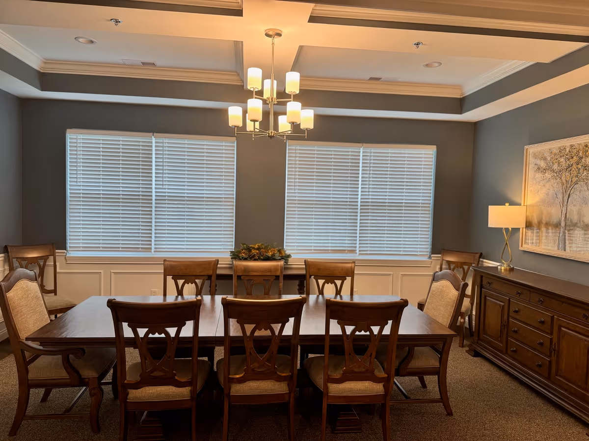 A dining room with a large wooden table surrounded by eight matching wooden chairs with cushioned seats. The room has two large windows with white blinds, a chandelier with multiple lights hanging from the ceiling, a sideboard with drawers and cabinets, a table lamp, and a framed painting of trees on the wall.